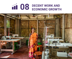 A woman from the project area stands confidently in front of sewing machines, representing the local women's collective that drives economic growth through sustainable livelihoods. This initiative, part of SDG 8: Decent Work and Economic Growth, includes activities such as mangrove honey collection and export, livestock support, and fisheries development to enhance household incomes in the region.