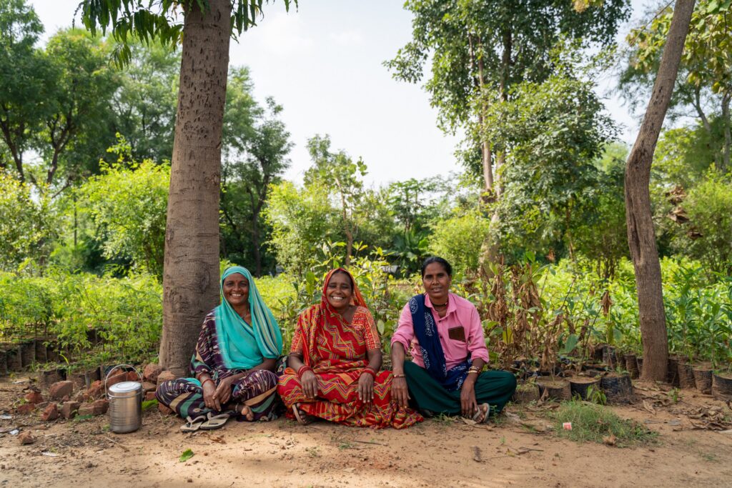 Three women from a farmer community group smiling, seated in front of a tree nursery